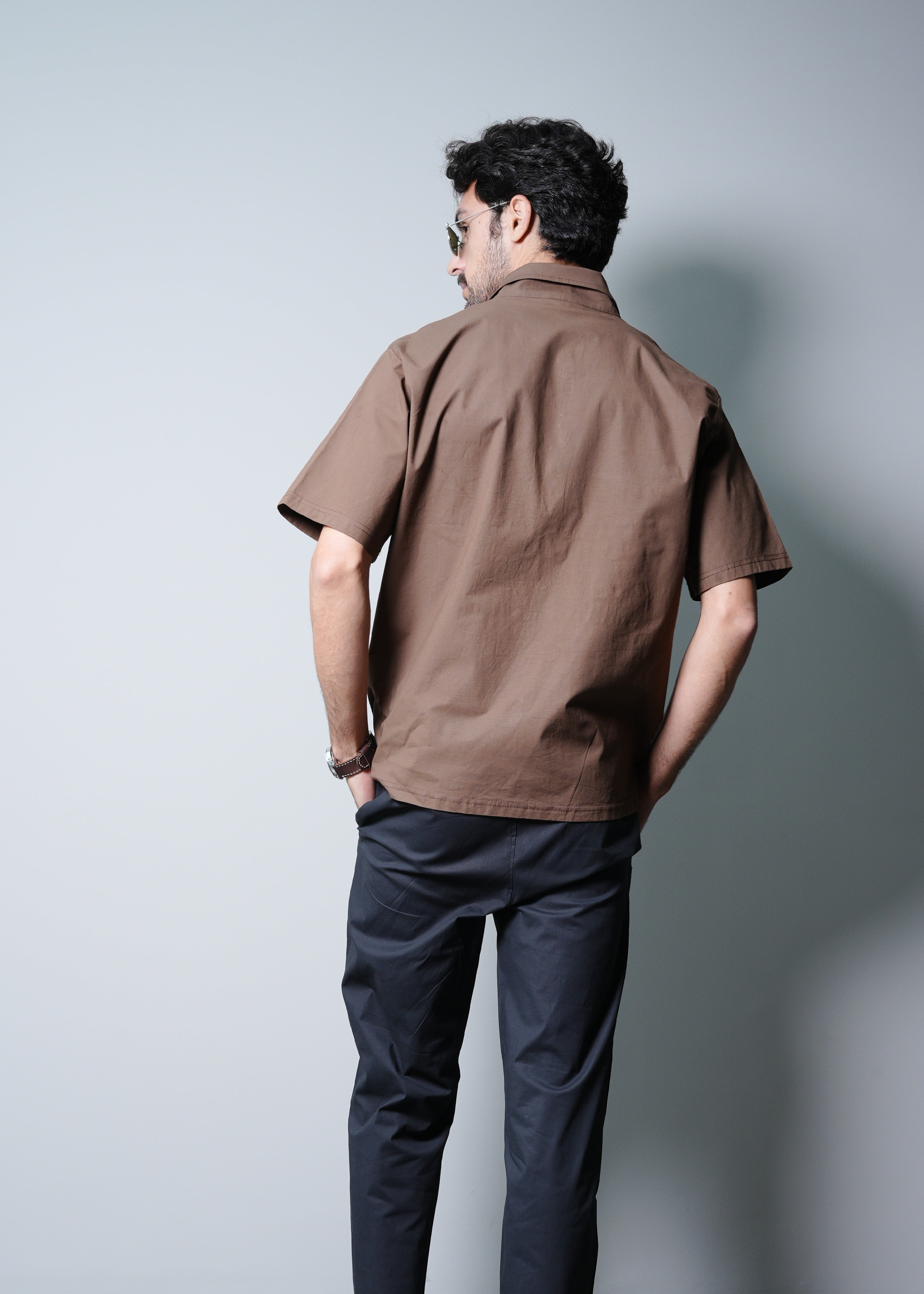 Man wearing a brown shirt and dark pants standing against a plain wall.